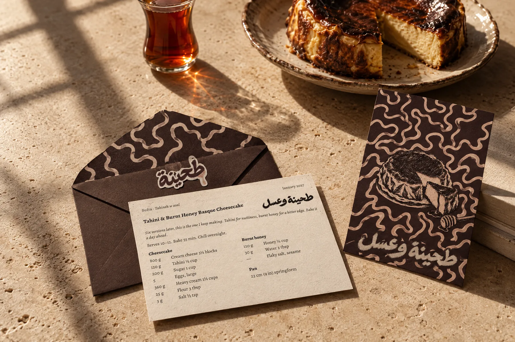 January's envelope opened on a travertine table in late sun, the recipe card resting on it, the postcard beside it, and the tahini and burnt honey Basque cheesecake behind with a glass of tea